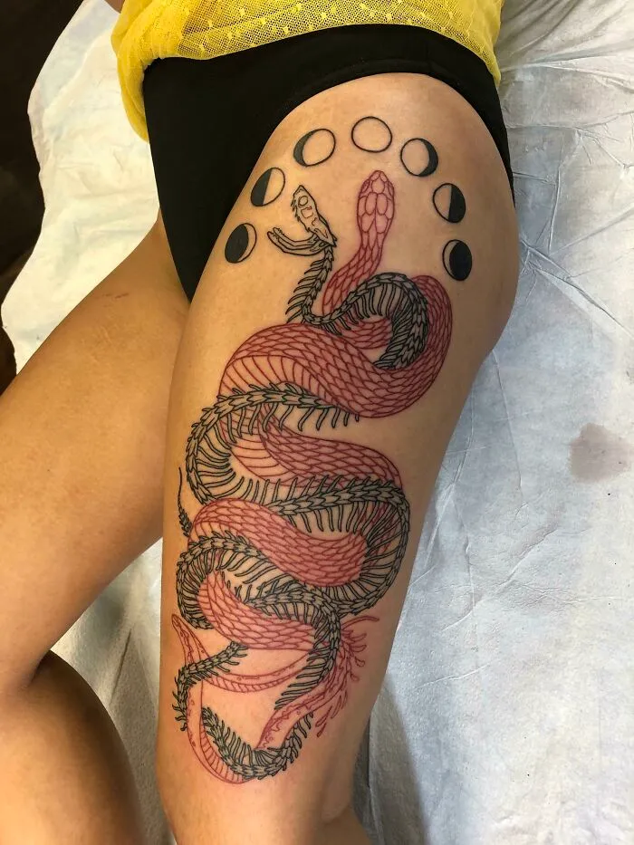Snake tattoo on a thigh with moon phases in black ink, showcasing intricate design and detail.