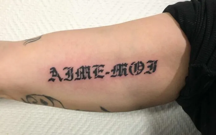 Gothic tattoo with "AIME-MOI" text in bold, black lettering on forearm.