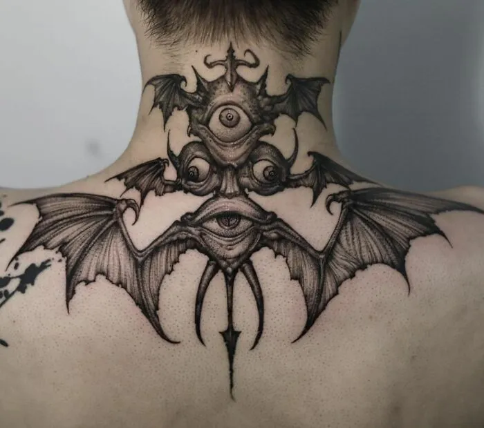 Gothic tattoo of a three-eyed creature with bat wings on the back of a person's neck and shoulders.