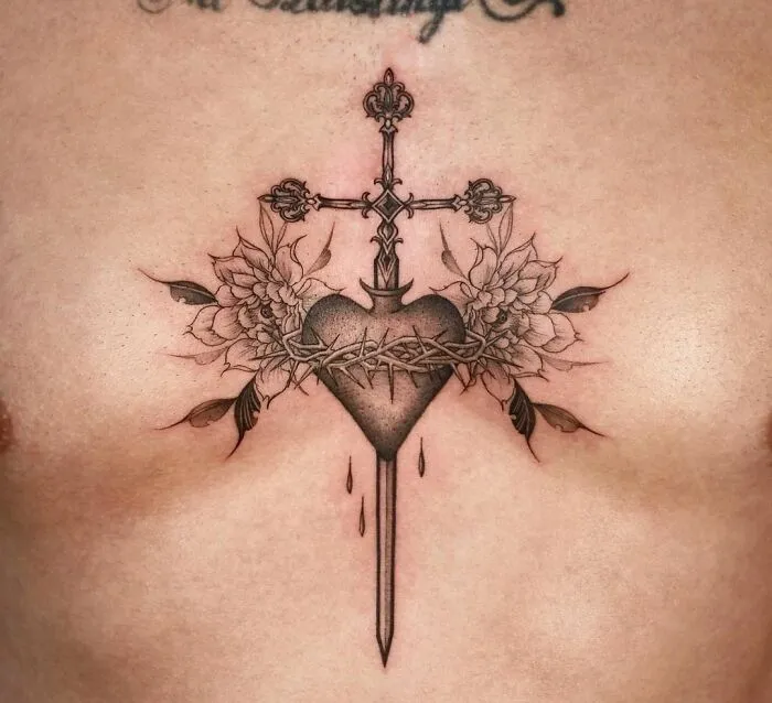 Gothic tattoo of heart pierced by a cross with floral details and barbed wire on chest.
