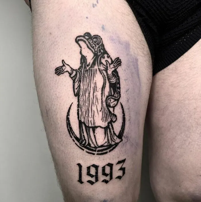 Gothic tattoo of a robed figure and the year 1993 inked on a person's thigh.