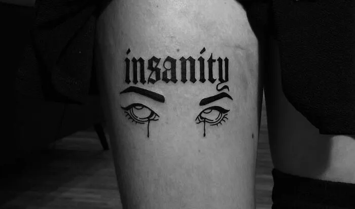 Gothic tattoo with word "insanity" above illustrated eyes on thigh.