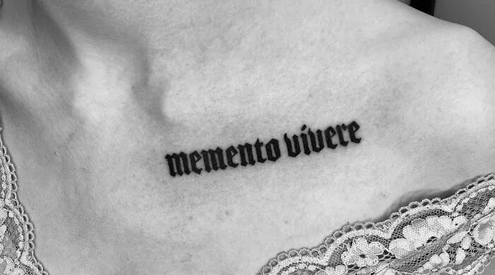 Gothic tattoo with "memento vivere" script on the collarbone in black ink.