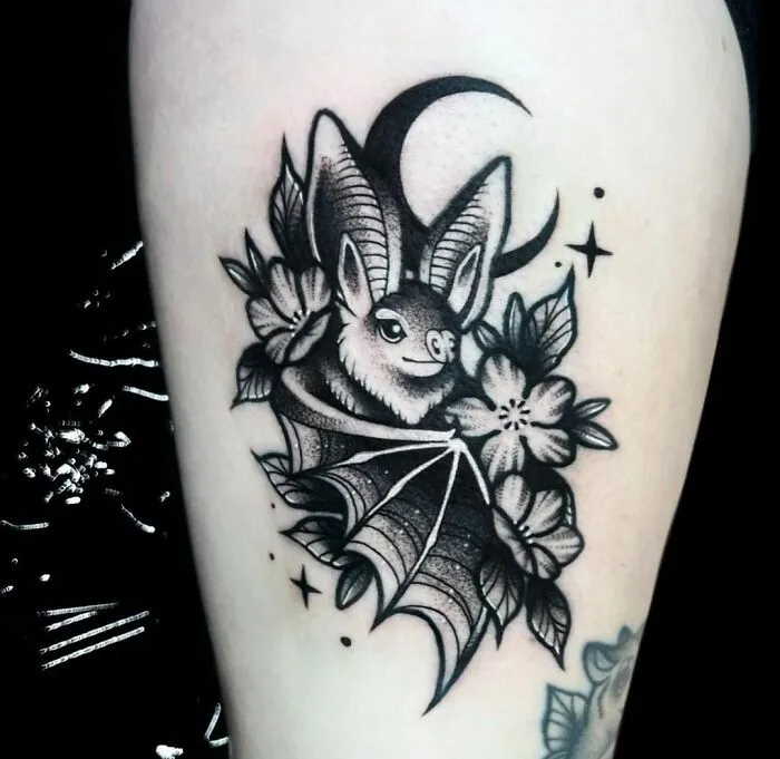 Gothic tattoo of a bat wrapped in flowers with a crescent moon and stars.