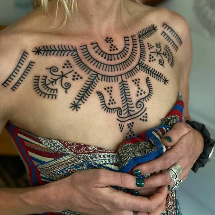 Woman displaying intricate tribal tattoo design on her chest, holding a colorful patterned fabric.