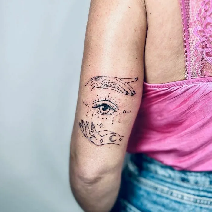 Arm tattoo featuring hands with an eye and celestial symbols, evoking yin yang duality in a pink top.