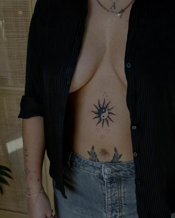 Person with a yin yang tattoo surrounded by sun rays on their abdomen, wearing a black shirt and jeans.