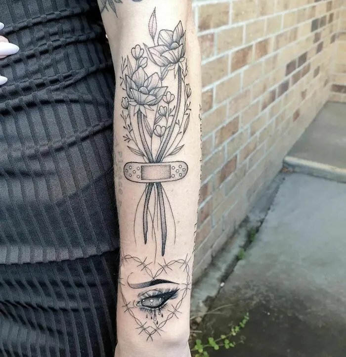 Arm tattoo featuring flowers wrapped in a band-aid, symbolizing mental health healing with a crying eye below.