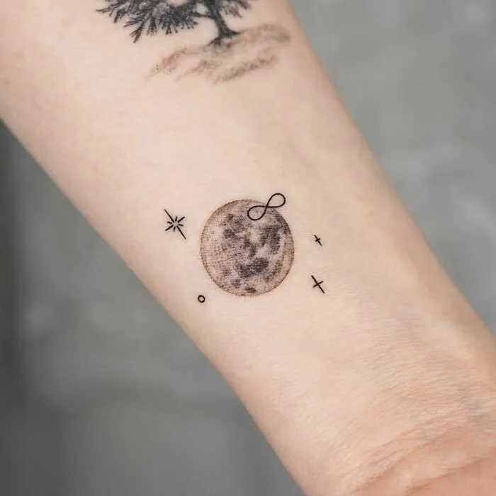 Infinity tattoo on wrist featuring a moon and small celestial symbols.