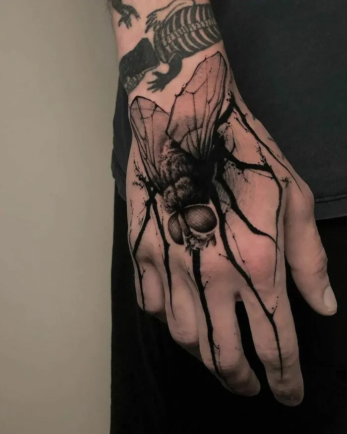 Hand with detailed gothic tattoo of a large fly and a skeletal design on the wrist.