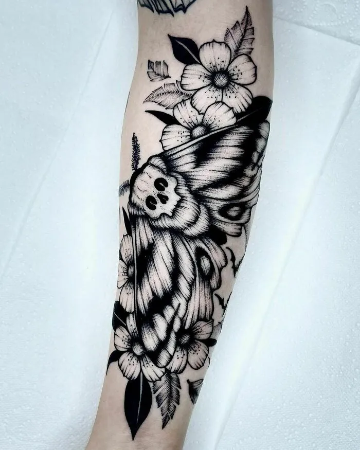 Gothic tattoo featuring a death moth and flowers on forearm, showcasing intricate black ink details.