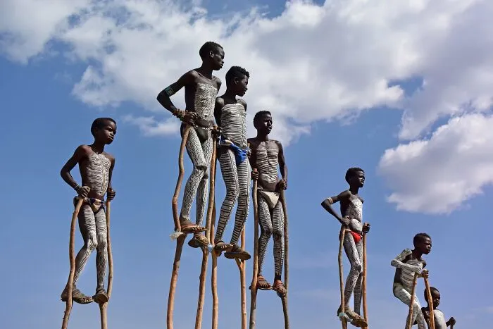 Banna Children In Ethiopia With Traditional Body Painting, Playing On Wooden Stilts By Wavrik