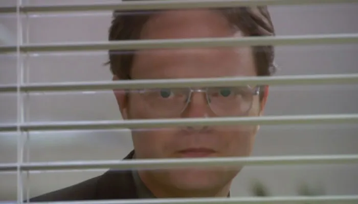 Dwight Schrute with a scary look on his face looking through blinds
