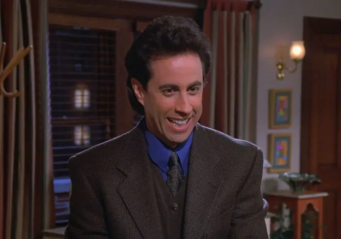 Jerry wearing a brown suit and blue shirt, smiling indoors, representing funniest Seinfeld quotes about everyday life.