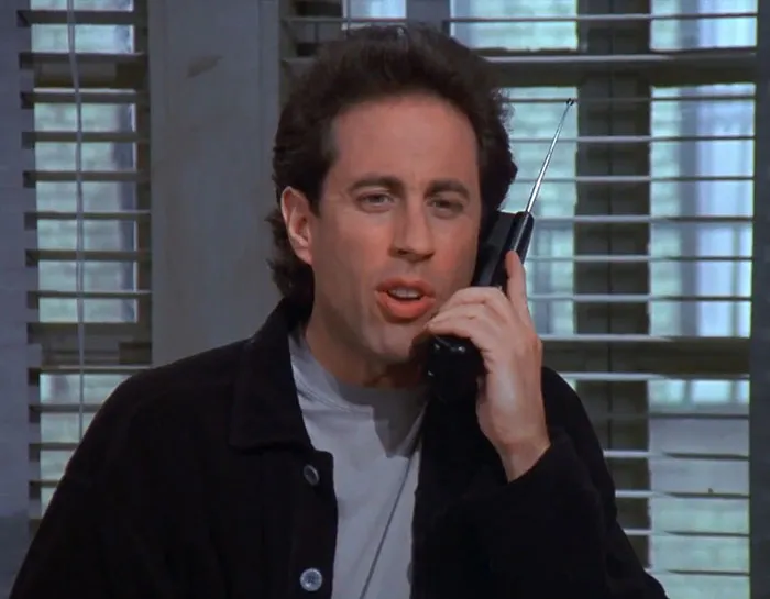 Jerry from Seinfeld wearing black jacket and gray shirt, speaking on a phone, illustrating funniest Seinfeld quotes on everyday life.