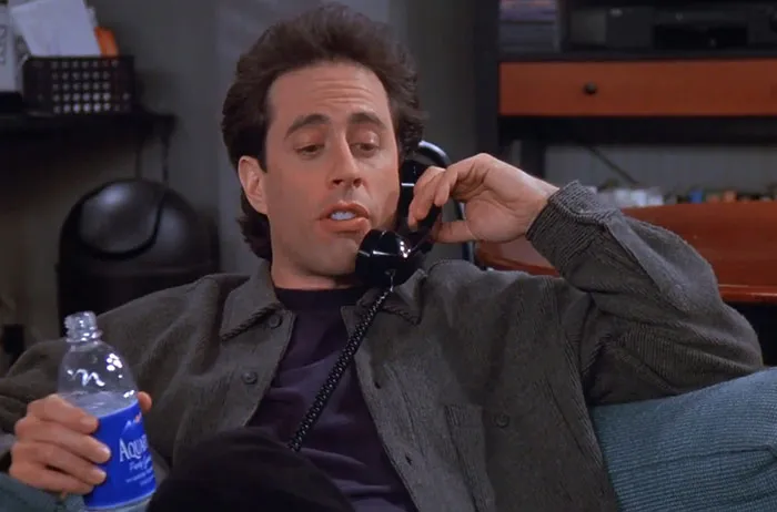 Jerry wearing gray sweater and dark blue shirt, talking on phone, a classic moment from funniest Seinfeld quotes about everyday life.
