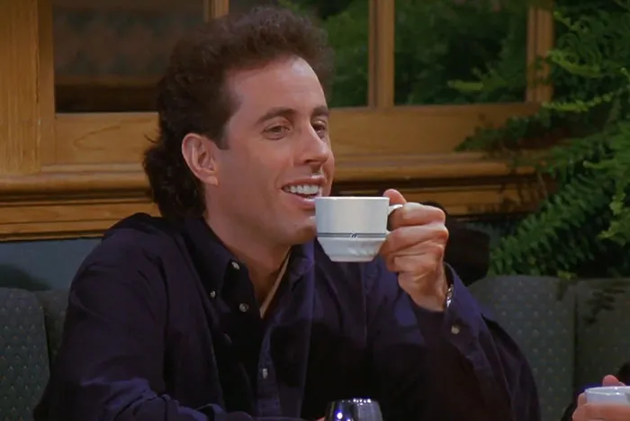 Jerry from Seinfeld wearing a blue shirt and drinking coffee, illustrating funniest Seinfeld quotes about everyday life.