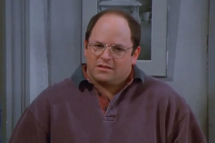 George wearing a purple sweater from Seinfeld, one of the funniest Seinfeld quotes moments that sum up everyday life.