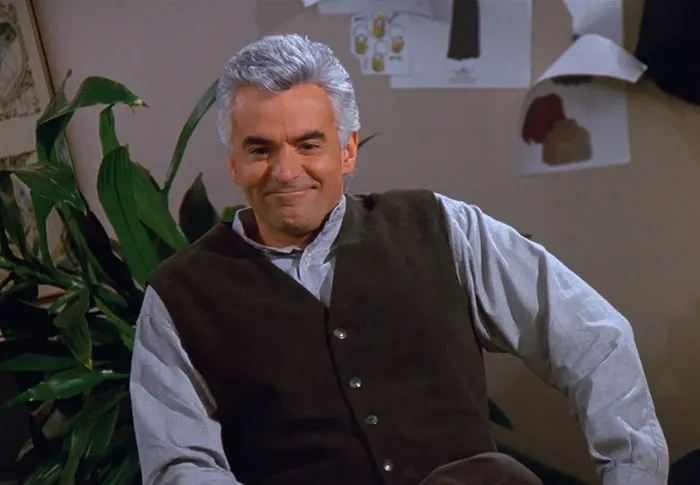 J. Peterman wearing brown vest and blue shirt, smiling in a casual setting, capturing funniest Seinfeld quotes vibe.