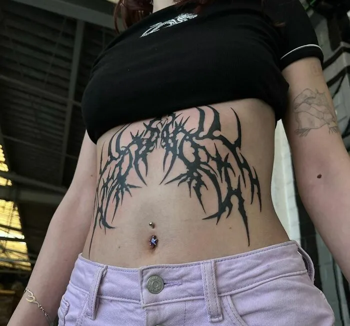 Woman with a tribal tattoo across her stomach, wearing a black top and pink pants, showcasing body art inspiration.