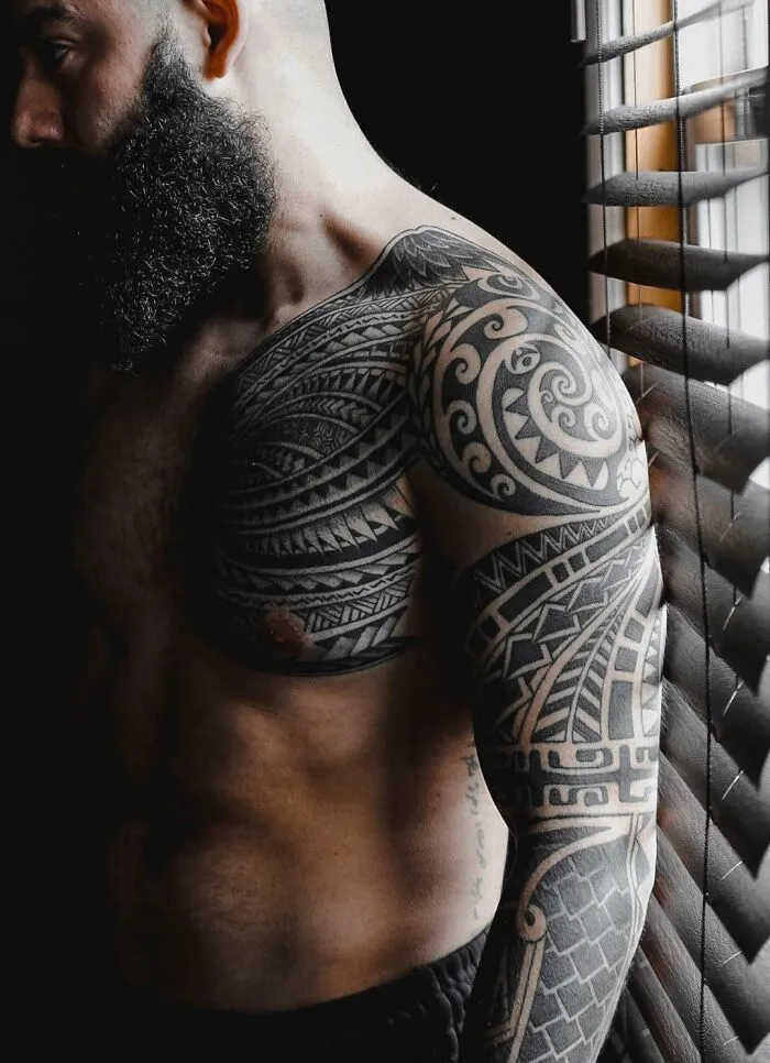 Man with intricate tribal tattoos on shoulder and arm, standing in soft light near window blinds.