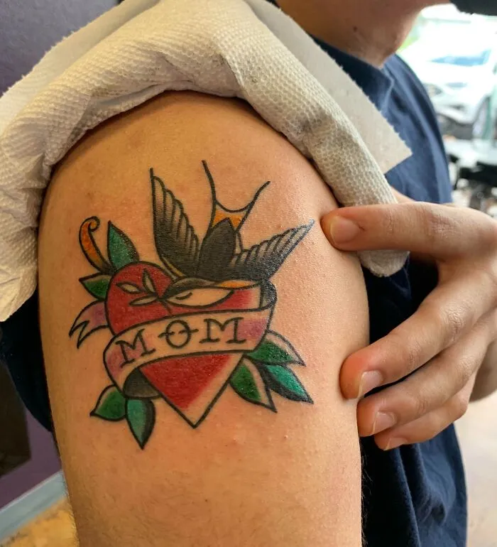 American traditional tattoo of a heart with the word "Mom" and a swallow on the upper arm.