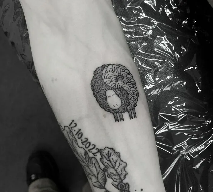 Black and gray sheep tattoo with date and leaves, symbolizing yin yang duality on forearm.