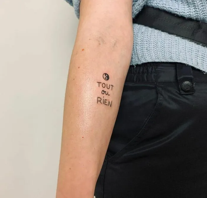 Yin Yang tattoo on arm with "Tout ou Rien" text, symbolizing duality.