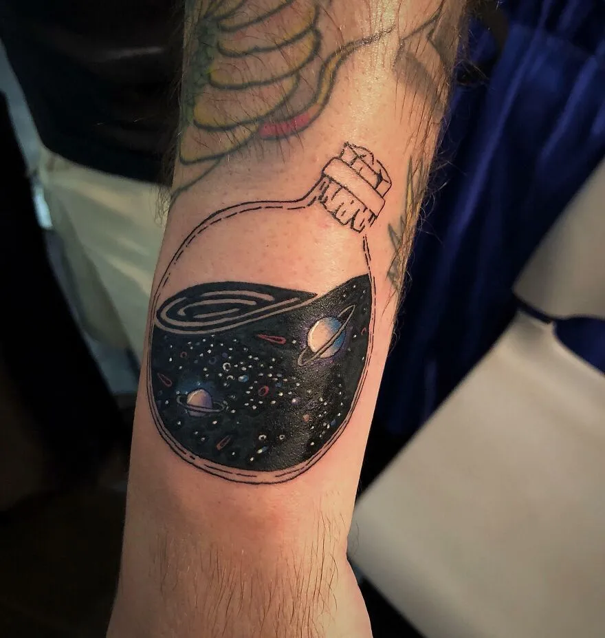 A space tattoo of a bottle holding a galaxy, featuring planets and stars, on a forearm.