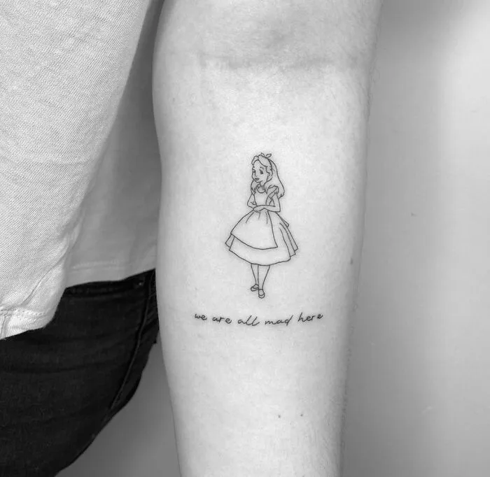 Cartoon tattoo showing a character and the phrase "we are all mad here" on an arm.