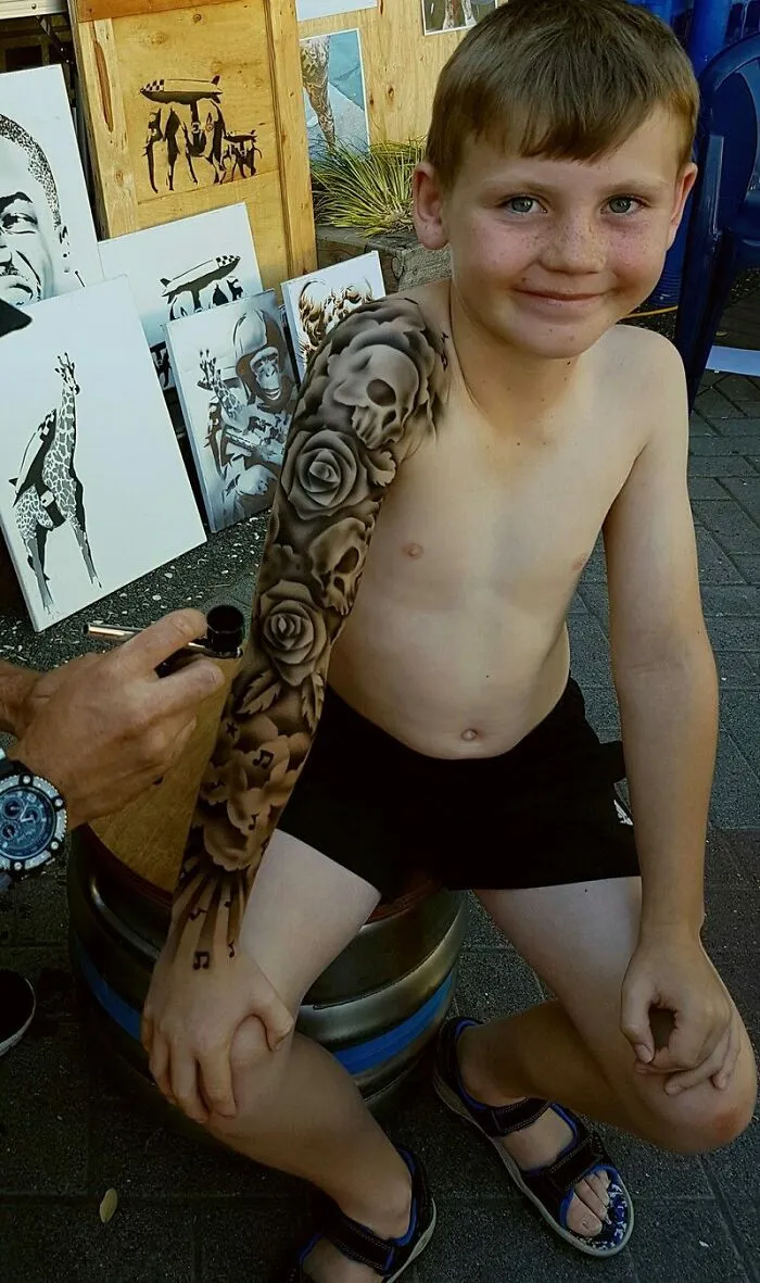 Hospitalized kid smiles with spray-on tattoo sleeve, featuring skulls and roses, applied by artist to brighten his day.