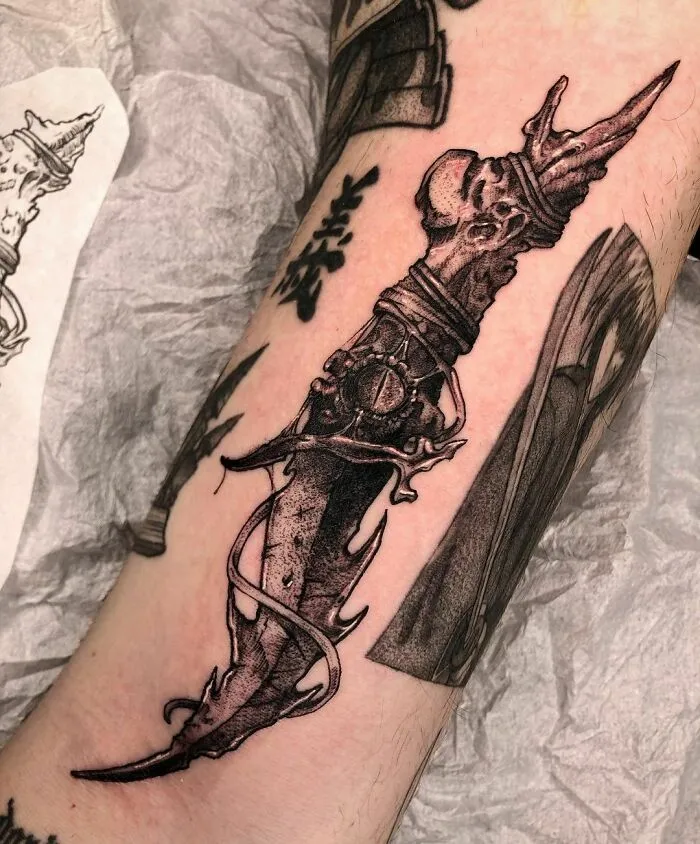 Gothic tattoo of a detailed ornamental dagger on an arm, featuring intricate dark elements and shading.