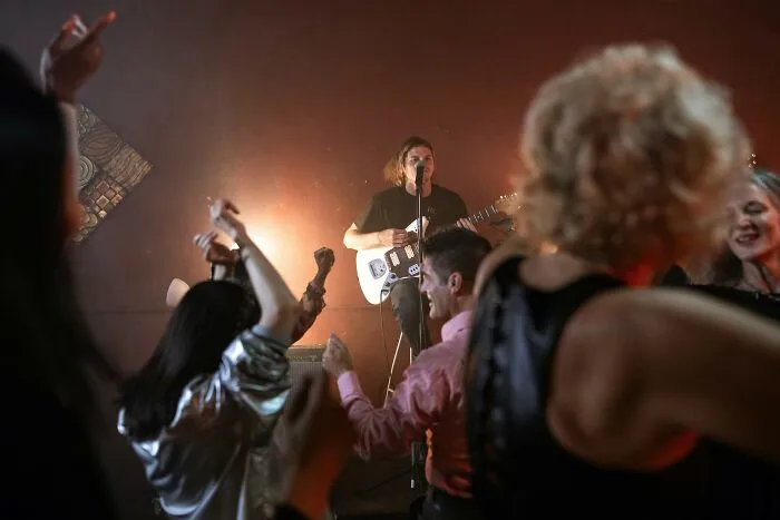 Person playing electric guitar on stage while people dance, capturing lively energy to impress friends with useless facts.