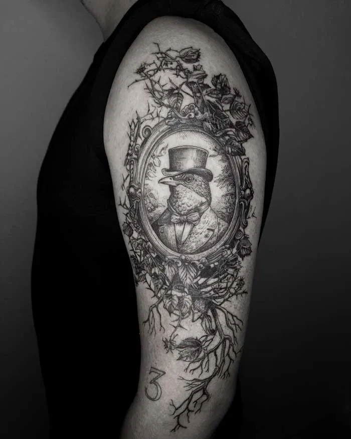 Gothic tattoo of a bird in a top hat on upper arm, intricate branches and frame detailing.