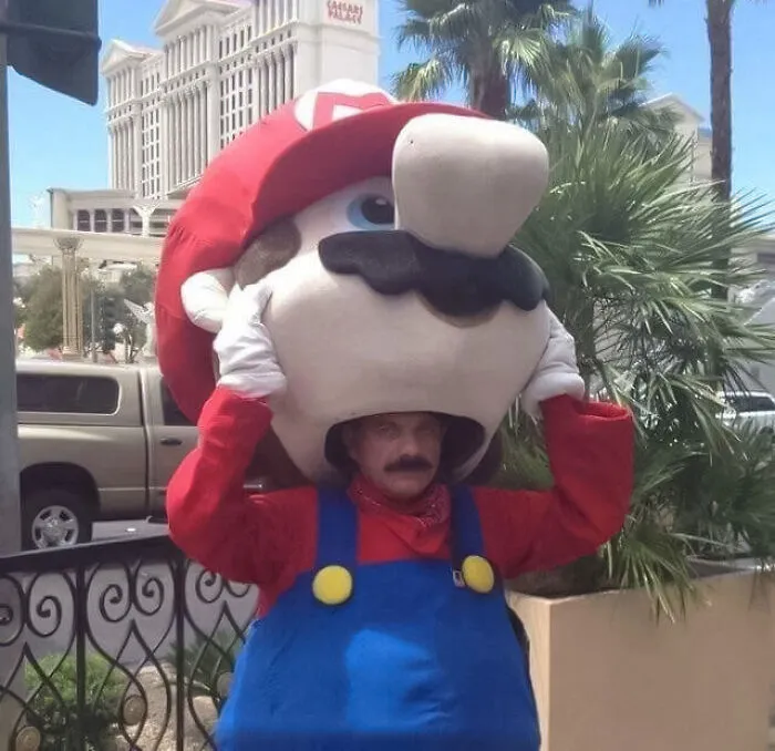 Person in oversized Mario costume head, adjusting it outdoors with a building and palm trees in the background.