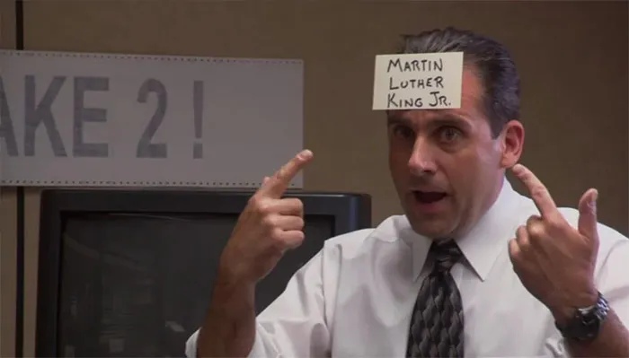 Michael Scott with a note on his forehead pointing at it, showcasing iconic boss moments in a casual office setting