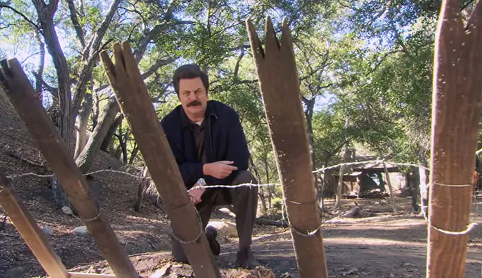 Ron Swanson crouching behind a wooden fence protecting his cabin in a forest setting, embodying rugged life wisdom.