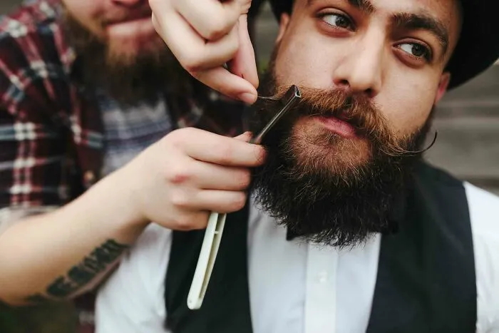 Barber shaving a bearded man with a straight razor in a vintage setting, representing useless facts to impress friends