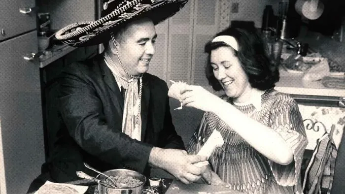 Glen Bell wearing a sombrero and sharing tacos with a woman, illustrating fun useless facts to impress friends.