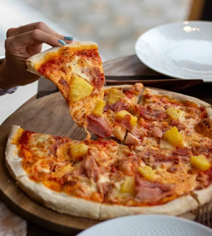 Person holding a slice of Hawaiian pizza on a wooden board, perfect for sharing fun useless facts with friends.