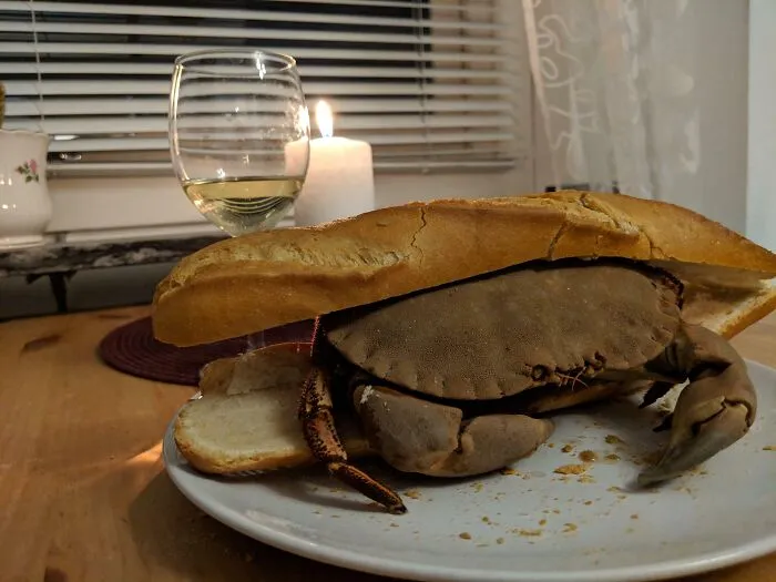 Cursed meme of a crab inside a sandwich roll on a plate, with a glass of wine and candle in the background.