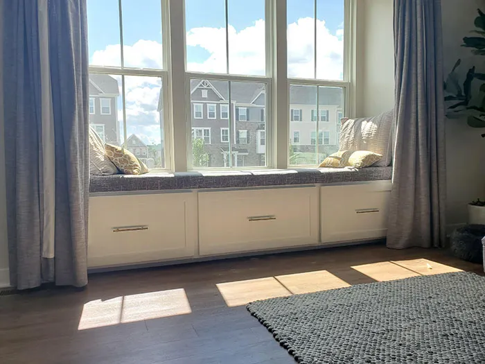 Monochromatic window seat with cushions and storage drawers under large bright windows in a cozy home space