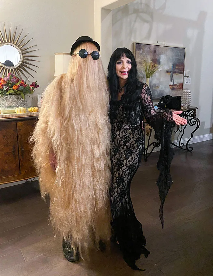 Two people in clever Halloween costumes, one with a long hair disguise and the other in a black lace dress indoors.