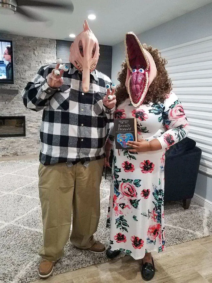 Couple wearing clever Halloween costumes with unique masks and props inside a living room setting.
