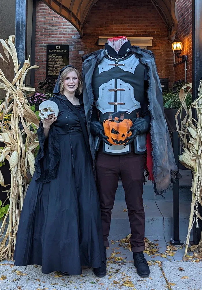 Woman in black dress holding skull stands next to headless man in clever Halloween costumes with pumpkin and armor.