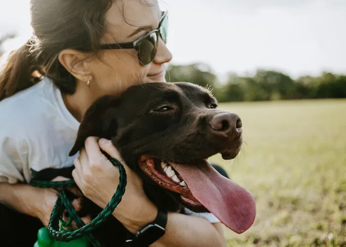 A woman in sunglasses hugging a happy brown dog outdoors, evoking a warm and thoughtful moment.