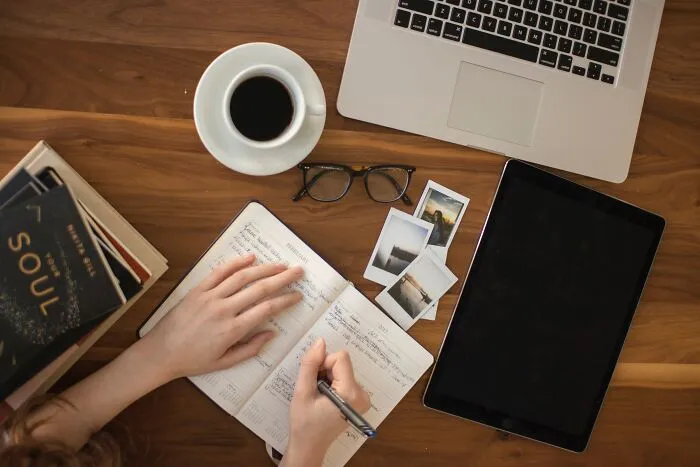 Person writing shower thoughts questions in notebook with laptop and coffee nearby.
