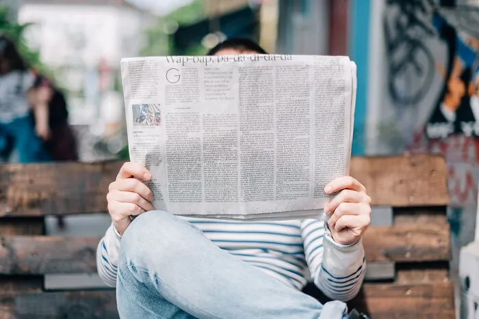 Person reading a newspaper on a wooden bench, pondering shower thoughts questions.