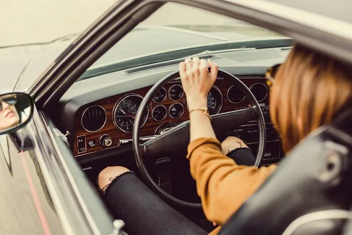 Person driving a vintage car, hand on the steering wheel, pondering shower thoughts questions quietly.
