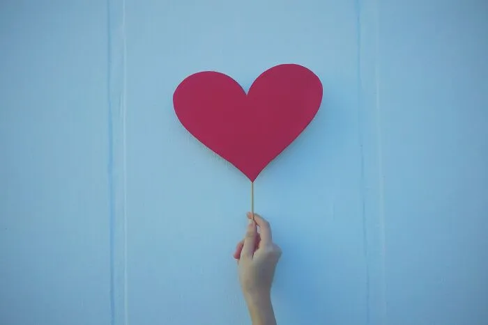 Hand holding a red heart-shaped cutout against a light blue background, symbolizing shower thoughts questions.